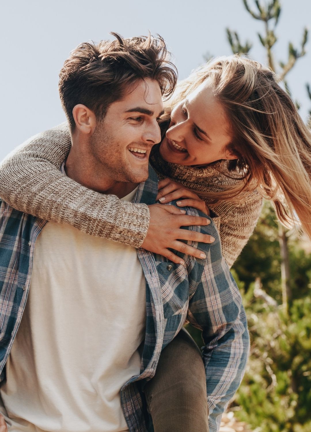 A woman gives a man a piggyback ride outdoors; both are smiling and dressed in casual clothing.
